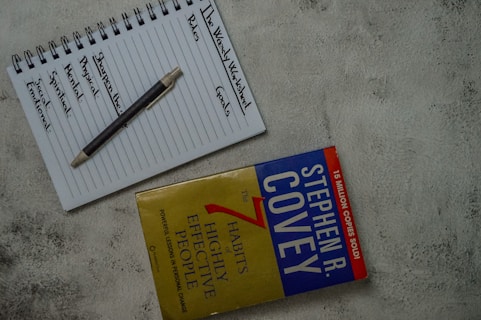 A top view of a notebook open to a page with the headings 'The Weekly Worksheet,' 'Roles,' and 'Goals' written at the top. Each column lists aspects like spiritual, physical, mental, and social/emotional, along with a central note 'Sharpen the Saw.' A pen rests diagonally across the page. Next to the notebook is the book 'The 7 Habits of Highly Effective People' by Stephen R. Covey, placed on a textured gray surface.