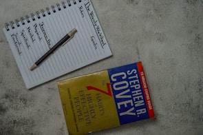 A top view of a notebook open to a page with the headings 'The Weekly Worksheet,' 'Roles,' and 'Goals' written at the top. Each column lists aspects like spiritual, physical, mental, and social/emotional, along with a central note 'Sharpen the Saw.' A pen rests diagonally across the page. Next to the notebook is the book 'The 7 Habits of Highly Effective People' by Stephen R. Covey, placed on a textured gray surface.