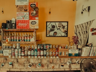 A cozy bar setup with a variety of bottled alcoholic beverages lined up on wooden shelves against a warm, orange wall. There are several branded boxes including Corona Extra, Budweiser, and Hoegaarden. Books and decor items are placed on the shelf, and there are fairy lights draped around a stone ledge. A wall-mounted speaker, decorative items, and a picture of cups and jars add to the homey atmosphere.