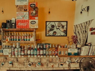 A cozy bar setup with a variety of bottled alcoholic beverages lined up on wooden shelves against a warm, orange wall. There are several branded boxes including Corona Extra, Budweiser, and Hoegaarden. Books and decor items are placed on the shelf, and there are fairy lights draped around a stone ledge. A wall-mounted speaker, decorative items, and a picture of cups and jars add to the homey atmosphere.