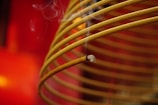 A close-up view of a spiral-shaped incense coil with smoke gently rising from its burning tip. The background is a vibrant red, adding a warm contrast to the brown hue of the incense coil.