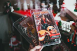 A hand is holding two packaged action figures against a background with Christmas decorations. The figure in the foreground is a muscular character wearing sunglasses, a red vest, and black pants, holding a large gun. The packaging includes a detailed illustration of the character. Behind this, another figure can be partially seen, dressed in boxing attire.