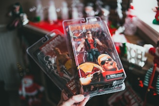 A hand is holding two packaged action figures against a background with Christmas decorations. The figure in the foreground is a muscular character wearing sunglasses, a red vest, and black pants, holding a large gun. The packaging includes a detailed illustration of the character. Behind this, another figure can be partially seen, dressed in boxing attire.