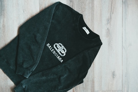 A dark-colored Balenciaga sweatshirt is laid out on a wooden floor. The design features a prominent logo in white at the center of the garment. The floor has a light wood grain texture, adding contrast to the dark fabric.