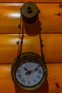 A vintage clock with Roman numerals is mounted on a wooden wall. The clock is encased in a rusty metallic holder, giving it an antique appearance. The background consists of polished wooden panels, enhancing the rustic aesthetic.