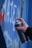 A hand is holding a spray paint can, applying graffiti to a blue surface. The graffiti includes white and red letters or symbols. The background appears to be metallic with some rust.