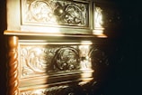 Intricate wood carvings on a piece of furniture with a swirling, floral design pattern. The surface is illuminated by warm sunlight, casting shadows and highlighting the texture of the wood. The handles are spherical and centered on the drawers.