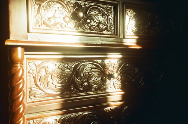 Intricate wood carvings on a piece of furniture with a swirling, floral design pattern. The surface is illuminated by warm sunlight, casting shadows and highlighting the texture of the wood. The handles are spherical and centered on the drawers.