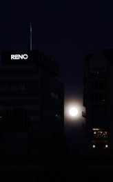 A bright full moon is visible between two dark, tall buildings at night. One of the buildings has 'RENO' lit up on it, and the moonlight casts a faint glow around its edges.