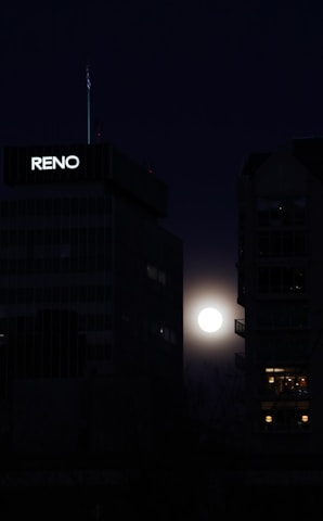 A bright full moon is visible between two dark, tall buildings at night. One of the buildings has 'RENO' lit up on it, and the moonlight casts a faint glow around its edges.