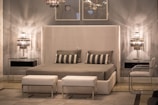 A modern bedroom features a large upholstered headboard with a neatly made bed covered in gray blankets and patterned pillows. The room is illuminated by two sleek, metallic table lamps on either side of the bed atop glass top nightstands. Above the bed, two minimalist framed artworks are hung on a gray wall. The decor is complemented by two white ottomans at the foot of the bed and a clear acrylic chair to the side.