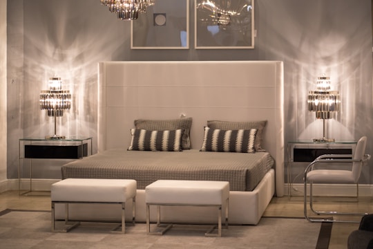 A modern bedroom features a large upholstered headboard with a neatly made bed covered in gray blankets and patterned pillows. The room is illuminated by two sleek, metallic table lamps on either side of the bed atop glass top nightstands. Above the bed, two minimalist framed artworks are hung on a gray wall. The decor is complemented by two white ottomans at the foot of the bed and a clear acrylic chair to the side.