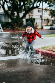 A child is joyfully jumping into a large puddle on a rainy street while wearing a red plaid jacket and a black beanie. Water splashes around their feet, capturing a moment of playfulness and movement. The background features urban elements like a sidewalk, trees, and residential buildings.