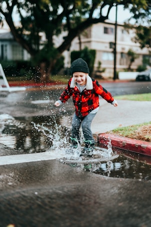 A child is joyfully jumping into a large puddle on a rainy street while wearing a red plaid jacket and a black beanie. Water splashes around their feet, capturing a moment of playfulness and movement. The background features urban elements like a sidewalk, trees, and residential buildings.