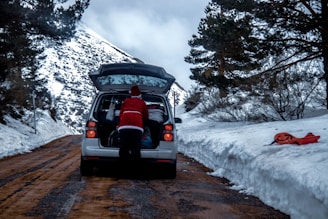 A person dressed in a Santa-like outfit is loading or unloading items from the back of a silver car parked on a snowy mountain road. The road is flanked by high snowbanks, and trees with snow are visible in the background. A red plastic sled lies on the snow to the right of the road.