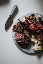 A platter filled with an assortment of food items including sliced salami, cheese cubes, crackers, nuts, and a wedge of cheese topped with pomegranate seeds. A knife is placed on the table near the plate.