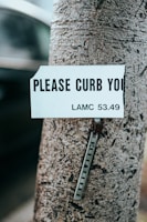 A close-up of a tree trunk with a damaged sign attached to it. The sign reads 'PLEASE CURB YOU' with a legislative reference 'LAMC 53.49' visible. The sign appears to be weathered and is missing part of the message. Below the sign, a metal strip with holes is fastened to the tree.