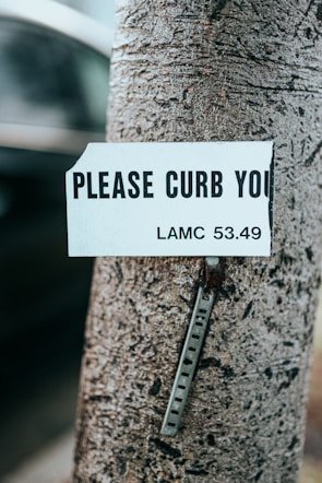 A close-up of a tree trunk with a damaged sign attached to it. The sign reads 'PLEASE CURB YOU' with a legislative reference 'LAMC 53.49' visible. The sign appears to be weathered and is missing part of the message. Below the sign, a metal strip with holes is fastened to the tree.