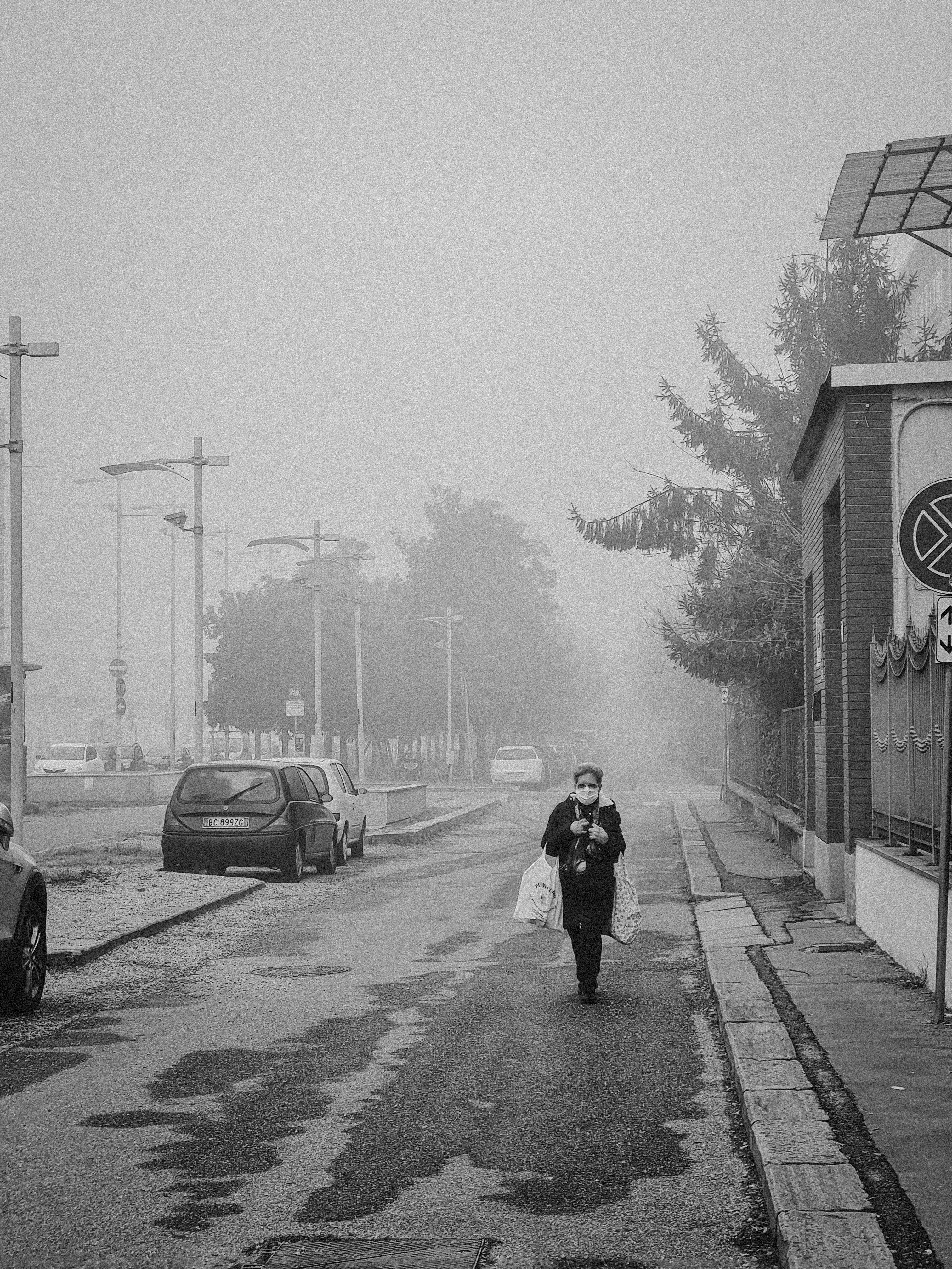 grayscale photo of man in black jacket and black pants walking on sidewalk