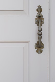 An ornate metal door handle with a traditional design is affixed to a cream-colored wooden door with paneled detail.