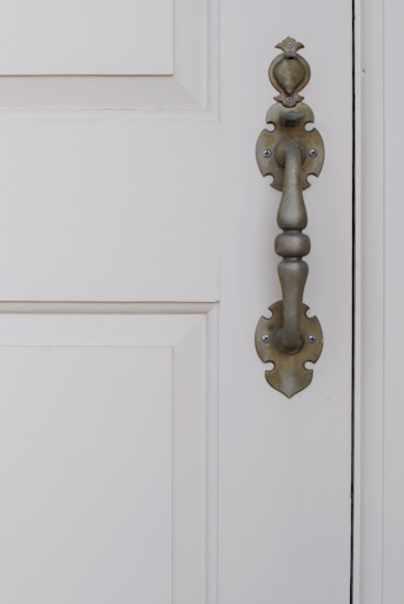 An ornate metal door handle with a traditional design is affixed to a cream-colored wooden door with paneled detail.
