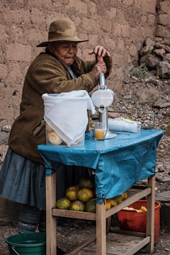 An elderly person wearing a hat and warm clothing is operating a manual citrus juicer on a makeshift stand. The stand is covered with a blue tarp and is laden with oranges or similar fruit. The setting appears rustic, with an earthy backdrop and rough stone or mud walls.