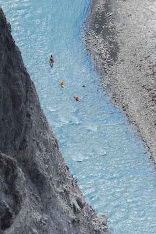 A narrow, winding river with clear blue water flows between rocky cliffs and a pebble-strewn shore. Three kayakers are paddling downstream, with vibrant kayaks in yellow, orange, and red adding contrast to the scene.