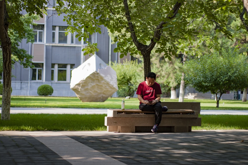 man in red shirt sitting on bench near tree during daytime