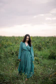 A model wearing a flowing bohemian dress in shades of teal, standing beneath a bright sunny sky.