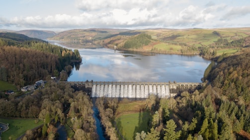 A large dam surrounded by a lush landscape of trees and hills, with a body of water stretching into the distance. The dam wall is releasing a cascade of water, and the scene is captured from an elevated perspective, highlighting the expansive natural scenery and the engineered structure.