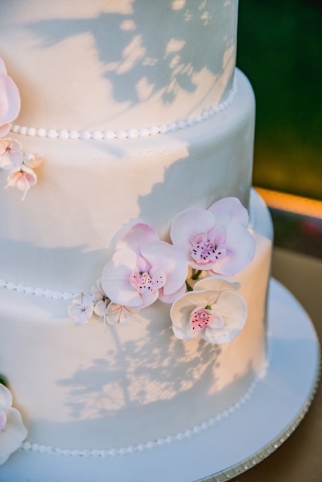 Elegant tiered cake adorned with delicate, pale pink and white sugar flowers, casting soft shadows on its smooth surface. The cake features subtle pearl-like decorations along the border of each tier.