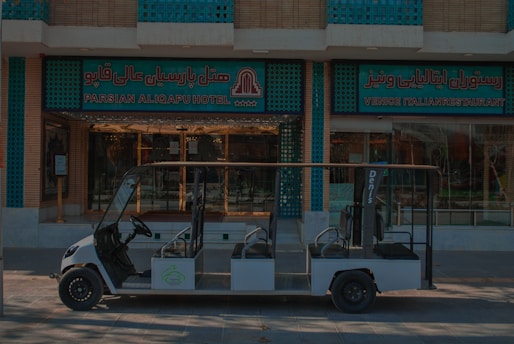 A small, open-sided electric vehicle is parked in front of a building with signs reading 'Parsian AliQapu Hotel' and 'Venice Italian Restaurant' in both English and Persian. The building features decorative turquoise and brickwork patterns.