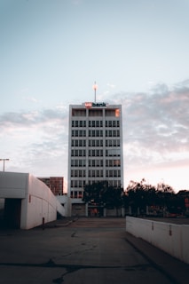 A tall office building with a sign on the top stands against a backdrop of a cloudy sky. The building is surrounded by trees and adjacent structures. The foreground includes a paved area with some cracks.