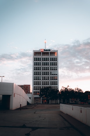 A tall office building with a sign on the top stands against a backdrop of a cloudy sky. The building is surrounded by trees and adjacent structures. The foreground includes a paved area with some cracks.