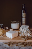 A wooden board on a table filled with a variety of cheeses, pasta, and a bottle of olive oil in the background. There is shredded cheese, a block of feta cheese, and crumbled blue cheese. A glass measuring cup with milk is also present along with a garlic clove.