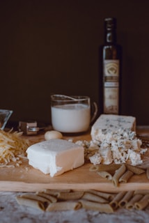 A wooden board on a table filled with a variety of cheeses, pasta, and a bottle of olive oil in the background. There is shredded cheese, a block of feta cheese, and crumbled blue cheese. A glass measuring cup with milk is also present along with a garlic clove.
