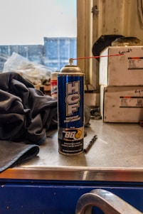 A can of waterproof spray lubricant labeled 'HCF' and manufactured by BG sits on a metal work surface. A red straw is attached to the nozzle, extending outward. Beside the can are industrial items including a fabric cloth and a small drill bit. The background features stacked boxes with indistinct writing, and a partially obstructed window revealing an industrial or workshop setting.