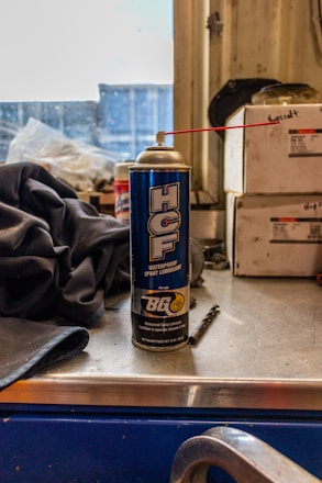 A can of waterproof spray lubricant labeled 'HCF' and manufactured by BG sits on a metal work surface. A red straw is attached to the nozzle, extending outward. Beside the can are industrial items including a fabric cloth and a small drill bit. The background features stacked boxes with indistinct writing, and a partially obstructed window revealing an industrial or workshop setting.