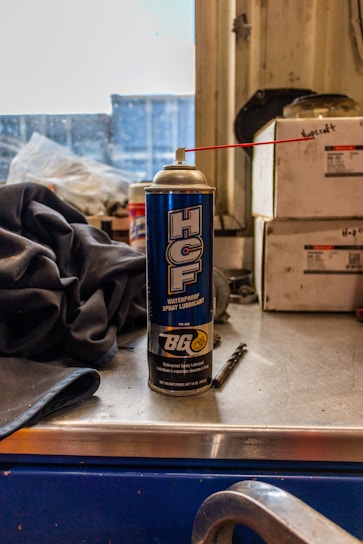 A can of waterproof spray lubricant labeled 'HCF' and manufactured by BG sits on a metal work surface. A red straw is attached to the nozzle, extending outward. Beside the can are industrial items including a fabric cloth and a small drill bit. The background features stacked boxes with indistinct writing, and a partially obstructed window revealing an industrial or workshop setting.