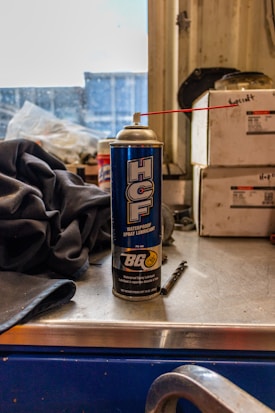 A can of waterproof spray lubricant labeled 'HCF' and manufactured by BG sits on a metal work surface. A red straw is attached to the nozzle, extending outward. Beside the can are industrial items including a fabric cloth and a small drill bit. The background features stacked boxes with indistinct writing, and a partially obstructed window revealing an industrial or workshop setting.