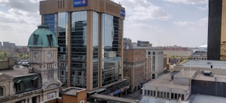 An urban scene featuring a mix of modern and historic architecture, including a tall, glass office building with a prominent BMO logo and a historic structure with a domed roof and clock tower. The street below is lined with various buildings, and the sky is partly cloudy.