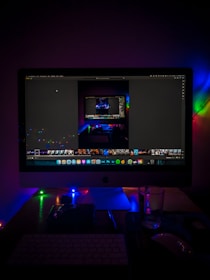 A desktop setup featuring a large monitor displaying an image editing software, surrounded by colorful LED lights. The scene includes a keyboard, a mouse, and a glass on the desk, creating a moody and tech-centric ambiance.