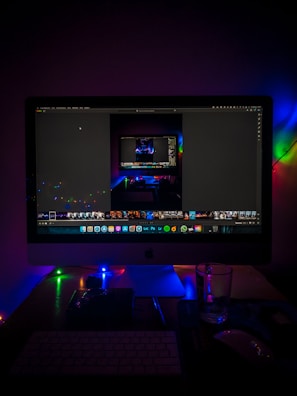 A desktop setup featuring a large monitor displaying an image editing software, surrounded by colorful LED lights. The scene includes a keyboard, a mouse, and a glass on the desk, creating a moody and tech-centric ambiance.