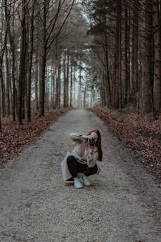 A person is crouching on a gravel path in a forest, holding a camera and taking a photograph. Tall trees line both sides of the path, creating a serene and natural tunnel effect. Fallen leaves cover the ground, adding to the autumnal atmosphere.
