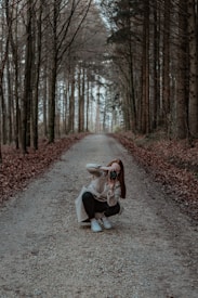 A person is crouching on a gravel path in a forest, holding a camera and taking a photograph. Tall trees line both sides of the path, creating a serene and natural tunnel effect. Fallen leaves cover the ground, adding to the autumnal atmosphere.