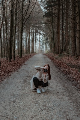 A person is crouching on a gravel path in a forest, holding a camera and taking a photograph. Tall trees line both sides of the path, creating a serene and natural tunnel effect. Fallen leaves cover the ground, adding to the autumnal atmosphere.