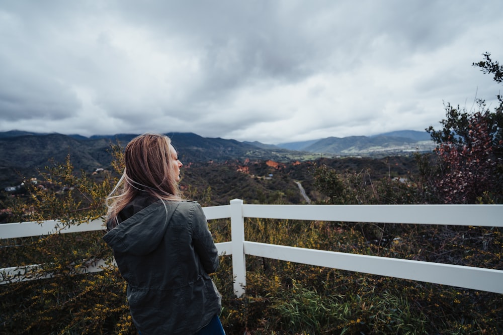 woman in blue denim jacket standing on green grass field near white wooden fence during daytime