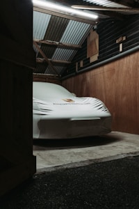 A car is parked inside a dimly lit garage, covered by a protective white cover with a logo on it. The garage structure features wooden walls and a corrugated metal roof, with fluorescent lighting overhead casting a soft glow.