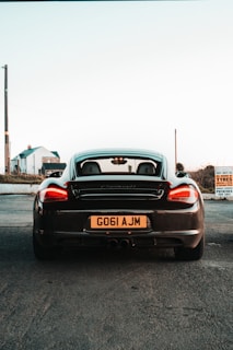 A black sports car seen from the rear, parked on a paved area with a visible license plate showing 'G061 AJM'. In the background, there are two buildings and a tall utility pole. A sign on the right indicates 'TYRES' and 'POTATOES FOR SALE'. The sky is clear with a pale hue, suggesting either dawn or dusk.