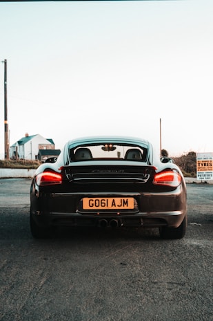 A black sports car seen from the rear, parked on a paved area with a visible license plate showing 'G061 AJM'. In the background, there are two buildings and a tall utility pole. A sign on the right indicates 'TYRES' and 'POTATOES FOR SALE'. The sky is clear with a pale hue, suggesting either dawn or dusk.